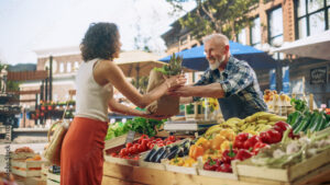 Cheerful Street Vendor Running a Small Farm Market Business, Selling Sustainable Fruits and Vegetables. Happy Middle Aged Man Filling a Recycled Paper Shopping Bag with Local Natural Food